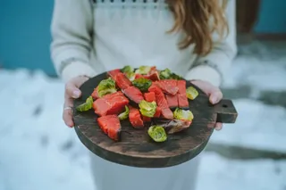 O persoană ține o plăcuță de lemn pe care sunt așezate bucăți de somon și legume verzi pe un fundal alb de zăpadă.