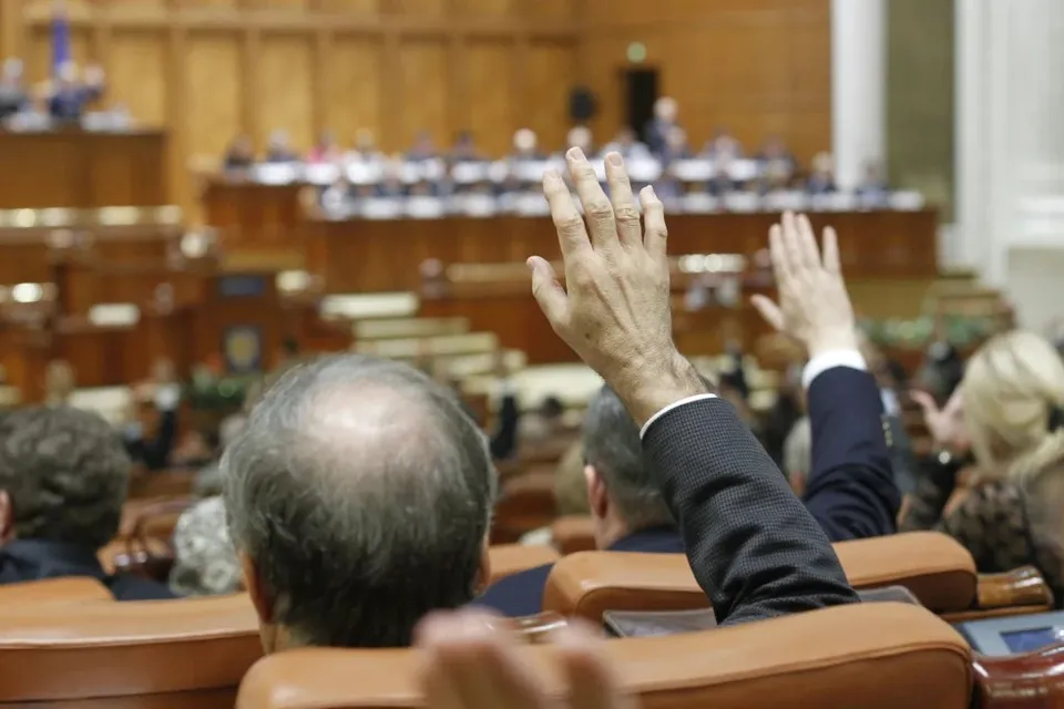 Două mâini ridicate deasupra unor persoane așezate în scaune maro într-o sală cu birouri de lemn și oameni în fundal.
