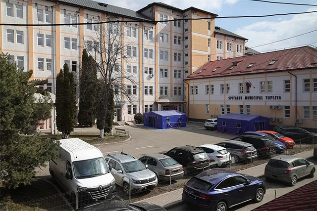 O clădire mare cu ferestre multiple este înconjurată de mașini parcate pe o suprafață asfaltată și de câțiva arbori înalți.