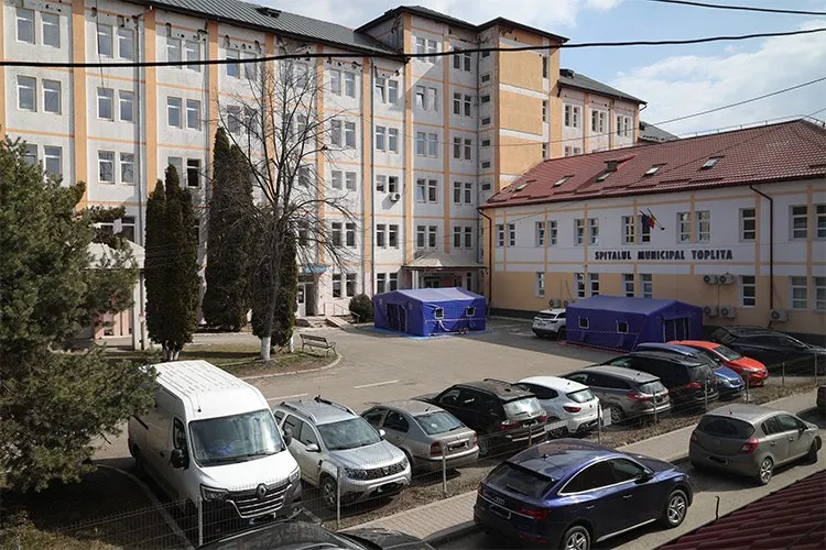 O clădire mare cu ferestre multiple este înconjurată de mașini parcate pe o suprafață asfaltată și de câțiva arbori înalți.