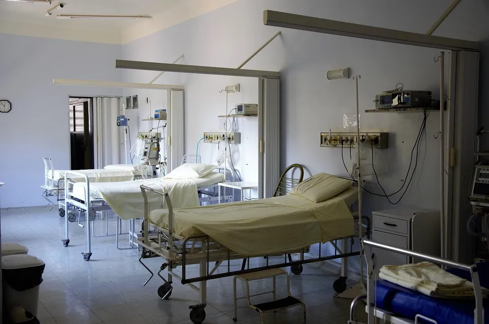 O cameră de spital conține mai multe paturi cu lenjerie albă, echipamente medicale și suporturi pentru perfuzii pe un fundal de pereți deschiși.