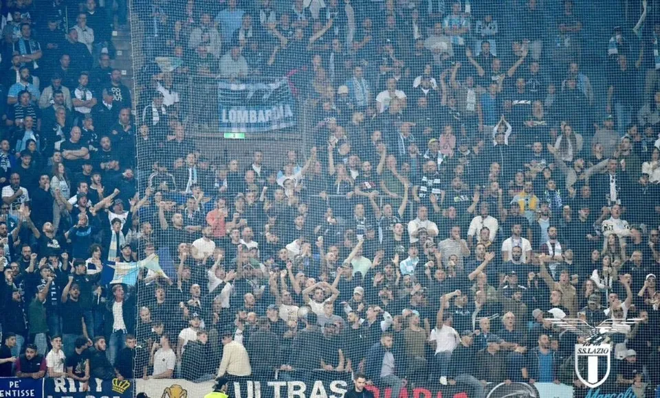 O mulțime de suporteri se adună în tribună, unii ridicând brațele și flând steaguri albastre și negre, într-un stadion de fotbal.