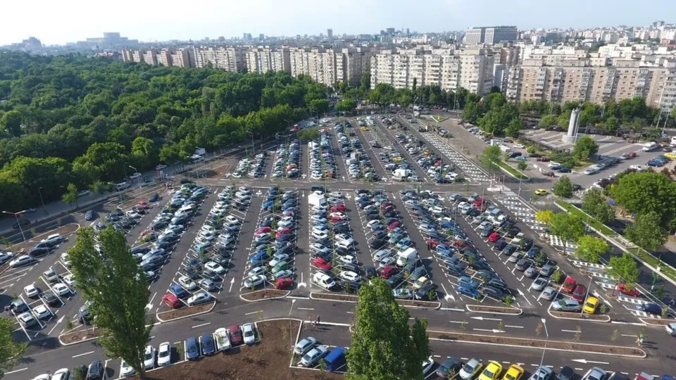 O vedere de sus asupra unui parc verde și a unei parcări mari pline de mașii colorate din diverse unghiuri.