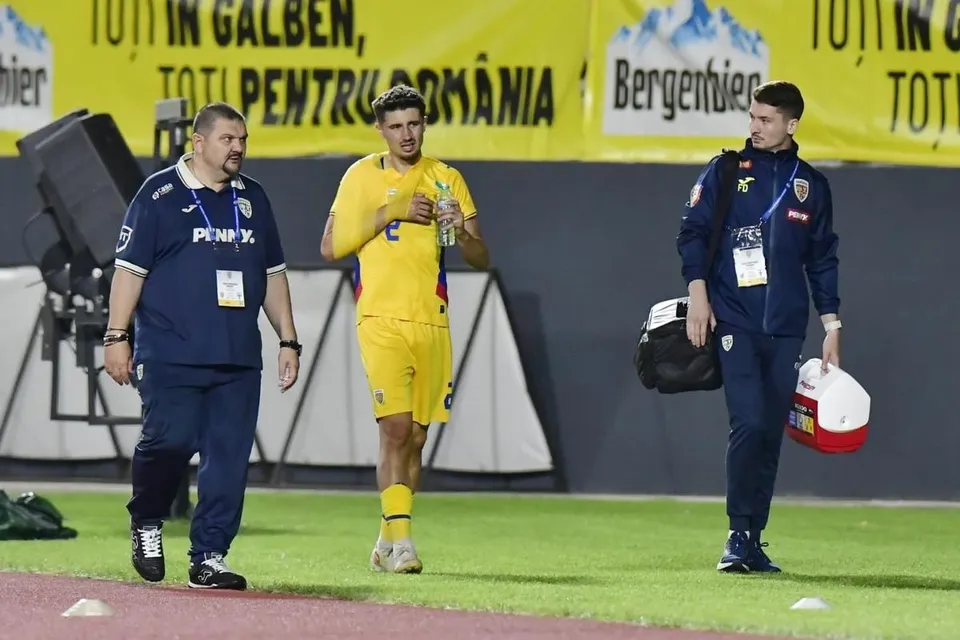 Un jucător de fotbal în echipament galben merge pe teren, însoțit de doi bărbați care poartă echipamente sportive și un sac medical.