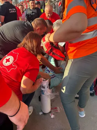 Mai mulți oameni în haine roșii și portocalii stau aplecați deasupra unei persoane, în timp ce un om în portocaliu ține o trusă medicală roșie.