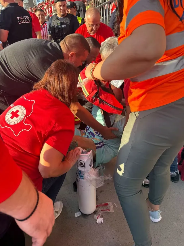 Mai mulți oameni în haine roșii și portocalii stau aplecați deasupra unei persoane, în timp ce un om în portocaliu ține o trusă medicală roșie.