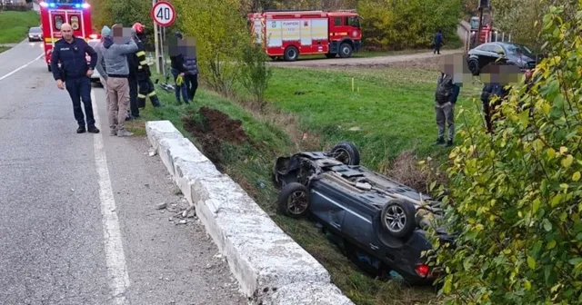 Un autoturism gri este răsturnat pe taluzul de lângă un cap de pod, lângă o stradă cu un camion de pompieri și persoane.