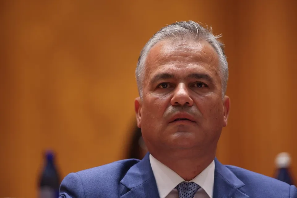 Un bărbat cu părul cărunt, purtând sacou albastru, cămașă albă și cravată gri, privește înainte într-un cadru de prim-plan.