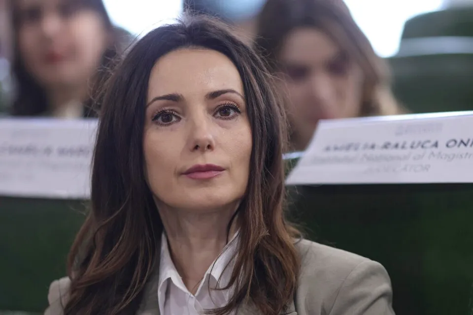 O femeie cu părul lung și brunet, purtând o cămașă albă și un sacou deschis, privește înainte într-o încăpere cu scaune verzi.