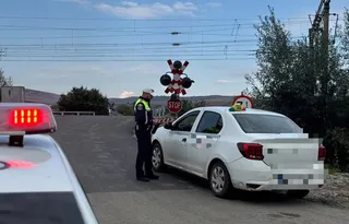 Un polițist stă lângă o mașină albă lângă o trecere la nivel cu semnalizare de tip barieră și semne de stop.