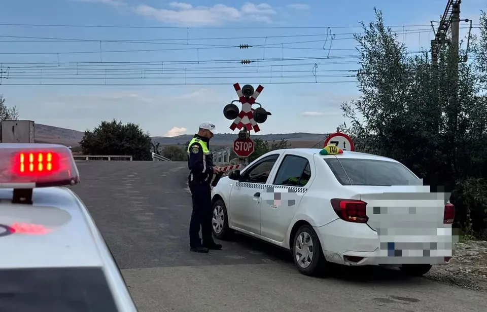 Un polițist stă lângă o mașină albă lângă o trecere la nivel cu semnalizare de tip barieră și semne de stop.