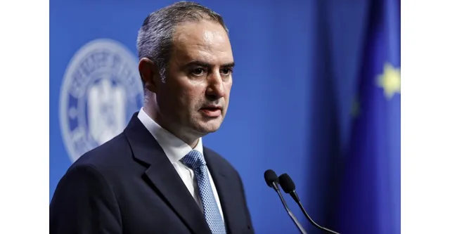 Un bărbat cu părul scurt, purtând sacou închis la culoare, cămașă albă și cravată albastră, stă în fața unui microfon pe un fundal albastru.