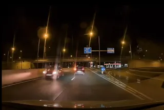 Două mașini circulă pe un drum asfaltat noaptea, sub iluminat public, lângă un pod și indicatoare rutiere suspendate.