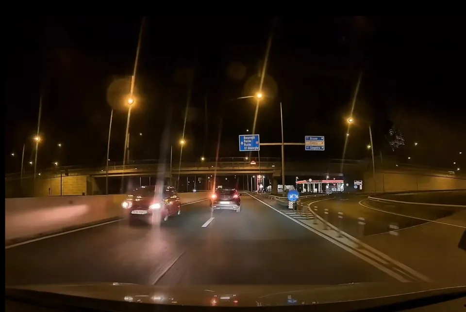 Două mașini circulă pe un drum asfaltat noaptea, sub iluminat public, lângă un pod și indicatoare rutiere suspendate.