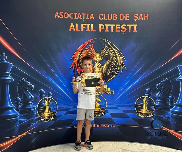 Un copil stă în picioare în fața unui panou decorativ cu tematică de șah, ținând o diplomă și un trofeu mic.