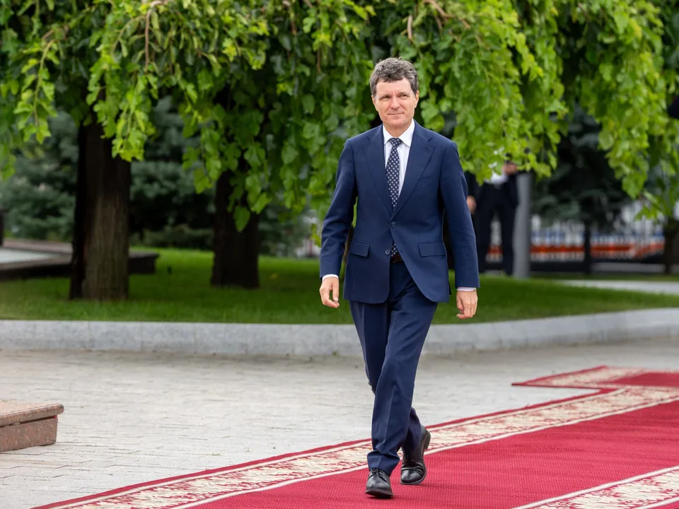 Un bărbat într-un costum albastru și cravată cu puncte merge pe un covor roșu aflat într-un spațiu exterior cu copaci verzi în fundal.