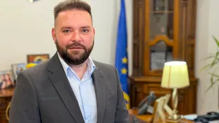 Un bărbat cu barbă și păr scurt, purtând sacou gri și cămașă albastră deschisă, stă în fața unui dulap de lemn și a unui steag.