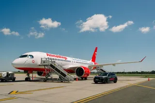 Un avion alb cu detalii roșii și textul Animawings stă pe o pistă de beton lângă o mașină neagră sub un cer cu nori albi.