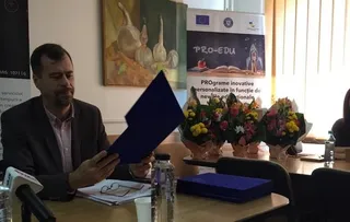 Un bărbat cu sacou și cămașă roz citește un dosar albastru la o masă cu flori și sticle de apă în fundal.