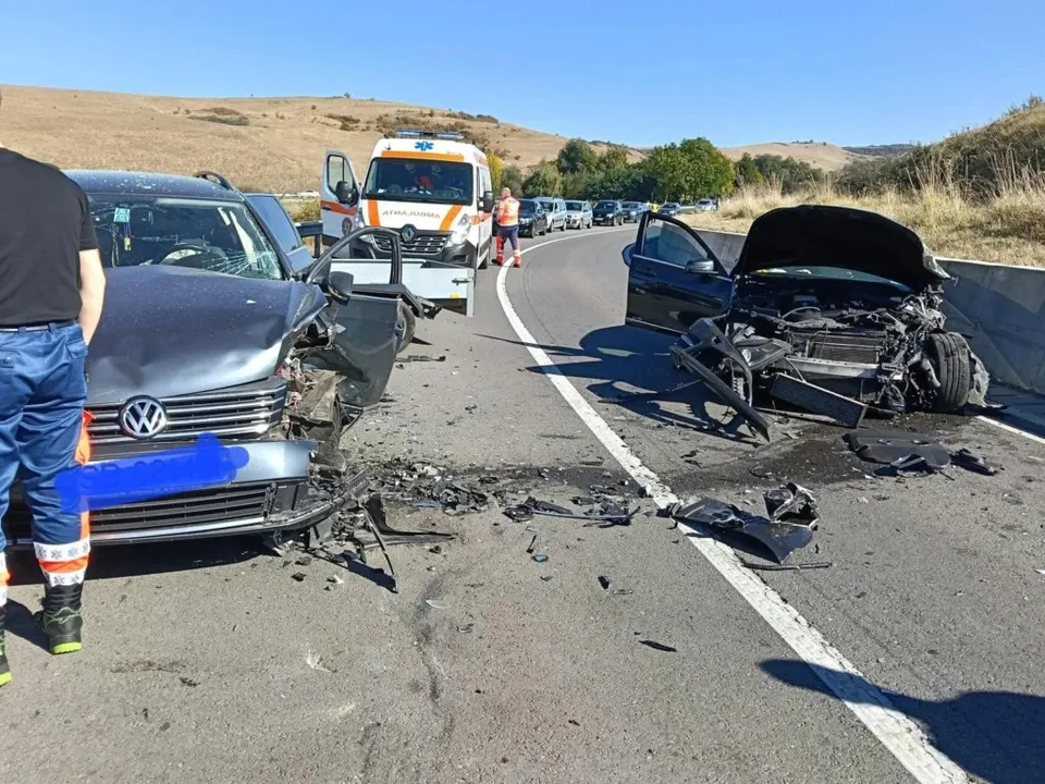 Două autoturisme accidentate stau pe un drum asfaltat lângă o ambulanță albă cu marcaje portocalii și persoane în uniformă.