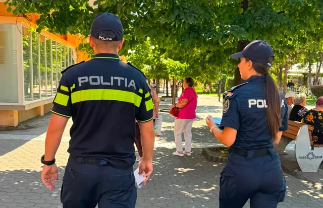 Doi polițiști în uniformă neagră cu detalii galben-verzui și inscripții sunt văzuți din spate pe un drum pietruit lângă copaci și oameni.