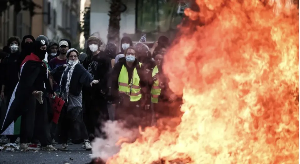 Un grup de persoane purtând măști și haine închise merge pe un drum lângă un flăcările mari și intense de foc.