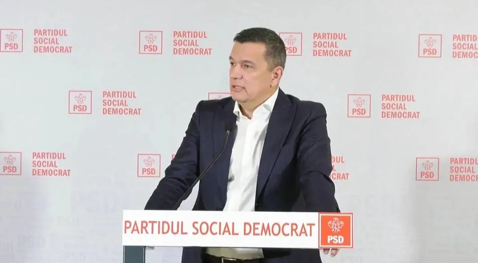 Un bărbat cu sacou închis și cămașă albă stă în spatele unui pupitru alb cu inscripții roșii în fața unui fundal alb cu logouri repetitive.