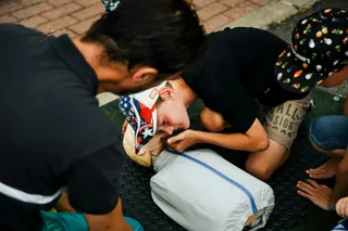 O persoană în cămașă neagră aplică manevre de prim ajutor unui individ întins pe o suprafață neagră texturată.
