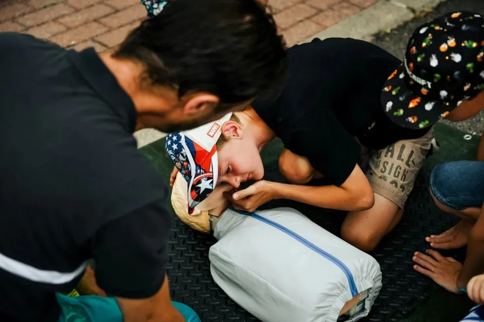 O persoană în cămașă neagră aplică manevre de prim ajutor unui individ întins pe o suprafață neagră texturată.