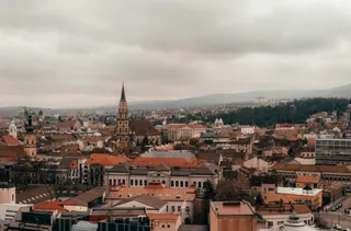 Vederea de sus asupra unui oraș cu numeroase clădiri cu acoperișuri colorate și un turn de biserică înalt sub un cer înnorat.