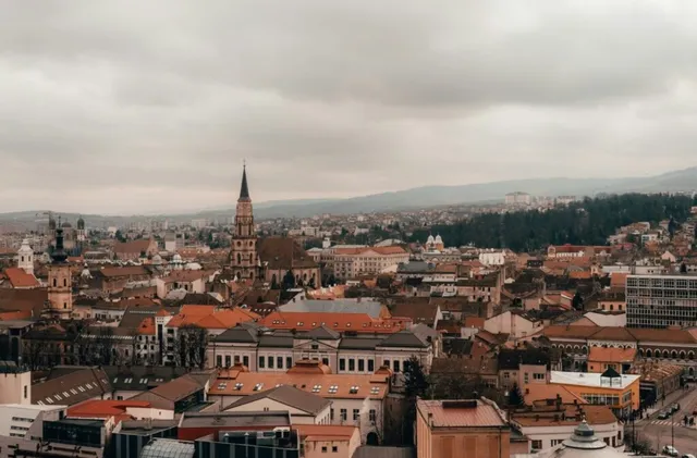 Vederea de sus asupra unui oraș cu numeroase clădiri cu acoperișuri colorate și un turn de biserică înalt sub un cer înnorat.