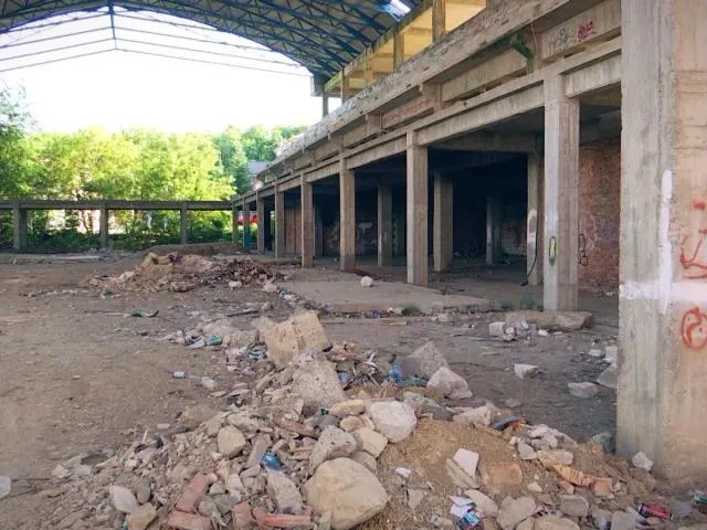 O structură de beton nefinisată cu coloane și grinzi deschise este acoperită de un acoperiș metalic, având resturi de pietre și deșeuri pe sol.