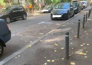 O serie de stâlpișori metalici gri sunt montați pe un trotuar asfaltat lângă mașini parcate pe o stradă urbană.