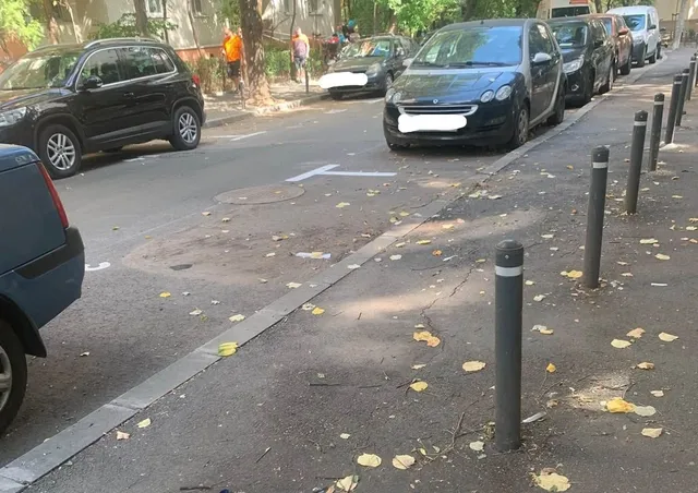 O serie de stâlpișori metalici gri sunt montați pe un trotuar asfaltat lângă mașini parcate pe o stradă urbană.