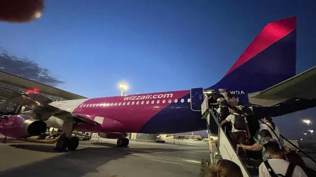 Un avion roz și alb este parcat pe o pistă de noapte, cu pasageri urcând pe scările de îmbarcare din partea din spate.