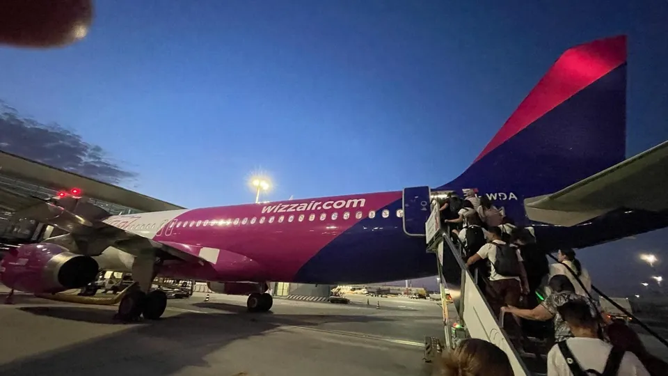 Un avion roz și alb este parcat pe o pistă de noapte, cu pasageri urcând pe scările de îmbarcare din partea din spate.