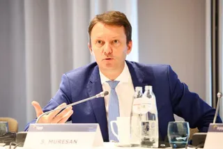 Un bărbat cu sacou albastru și cămașă albă cu cravată albastră stă la o masă cu microfon, sticle de apă și un pahar.