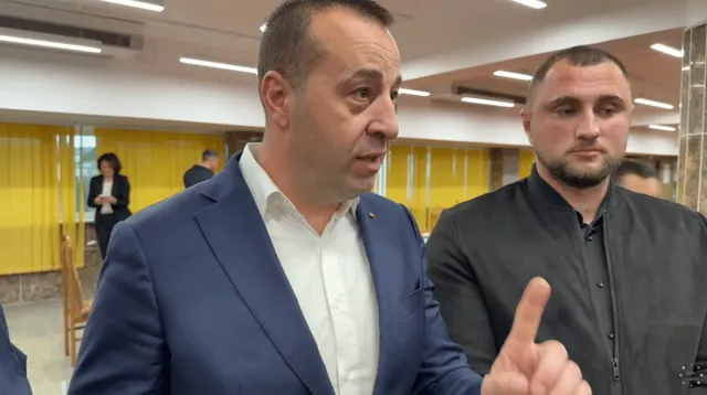 Un bărbat într-un sacou albastru și cămașă albă ridică degetul arătător, fiind însoțit de un alt bărbat într-o cămașă neagră.