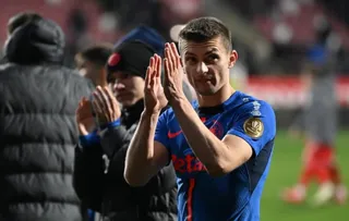 Un bărbat într-un tricou albastru aplaudă cu ambele mâini pe un teren de fotbal, în timp ce alți bărbați stau în spatele lui.