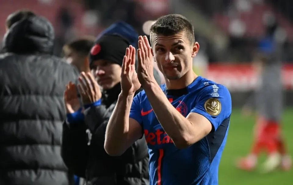 Un bărbat într-un tricou albastru aplaudă cu ambele mâini pe un teren de fotbal, în timp ce alți bărbați stau în spatele lui.