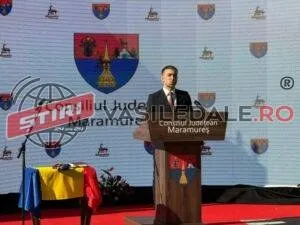 Un bărbat într-un costum închis la culoare stă în spatele unui podium de lemn în fața unui panou cu diverse embleme colorate.
