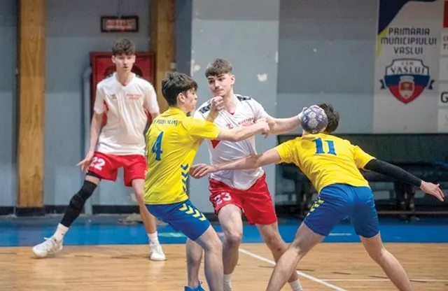 Patru tineri jucători de handbal în echipamente alb-roșii și galben-albastre participă la un meci pe un teren de interior.