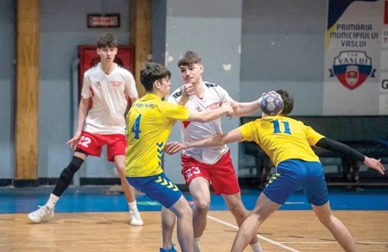 Patru tineri jucători de handbal în echipamente alb-roșii și galben-albastre participă la un meci pe un teren de interior.