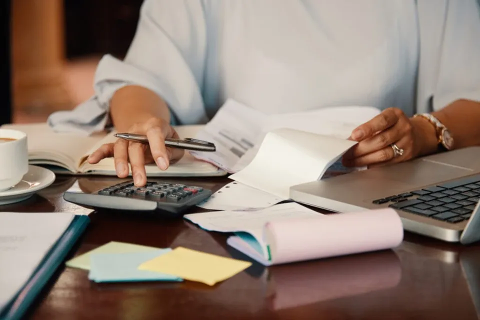 O persoană cu mâinile pe o masă folosește un calculator, un stilou și diverse hârtii lângă un laptop și o cană de cafea.
