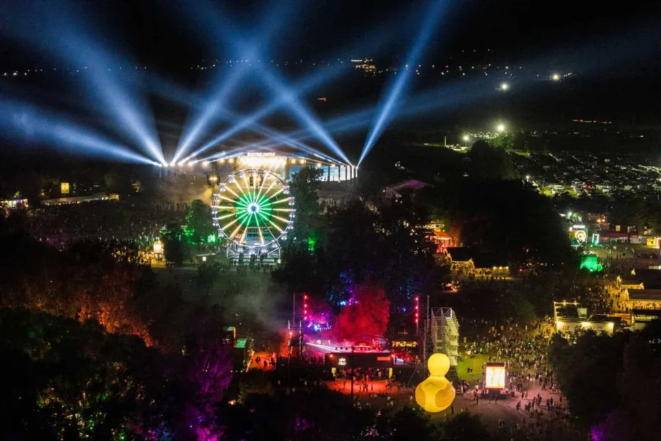 O roată feroviară iluminată și mai multe scene de festival cu lumini colorate și proiectoare puternice sub un cer de noapte.