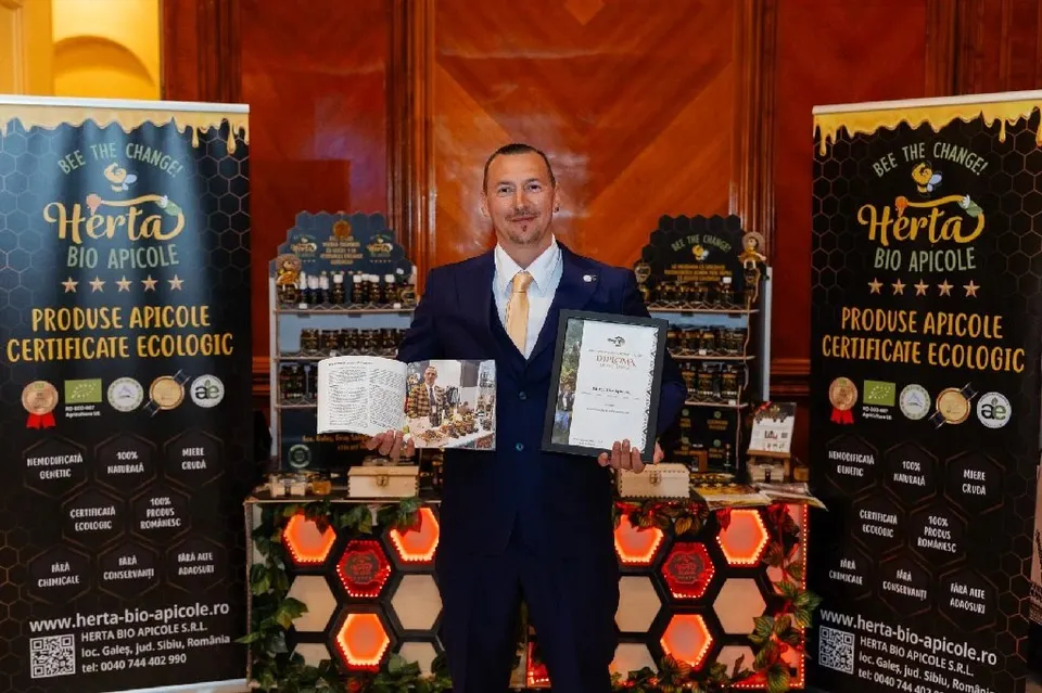 Un bărbat într-un costum albastru ține un certificat și o broșură în fața unor standuri cu produse apicole și panouri informative.