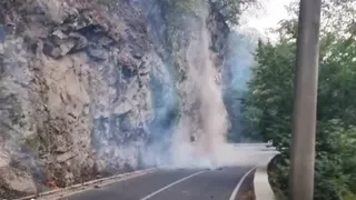 Drum asfaltat curbat trece pe lângă un versant stâncos, unde fum și flăcări mici se văd lângă șosea.