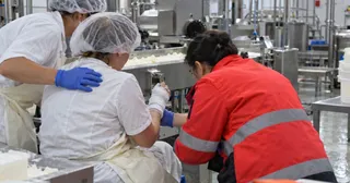 Trei persoane, dintre care două poartă haine albe și o persoană poartă o jachetă roșie, stau lângă un utilaj metalic într-un spațiu industrial.