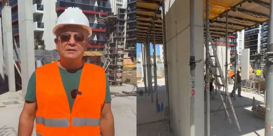 Un bărbat cu cască albă și vestă reflectorizantă portocalie stă în fața unor clădiri în construcție, lângă un șantier cu schele metalice.