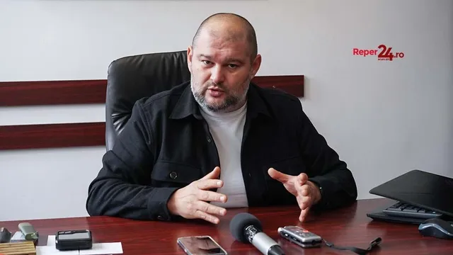 Un bărbat cu barbă și haină neagră stă la o masă de lemn în fața unui microfon și a unor dispozitive electronice.